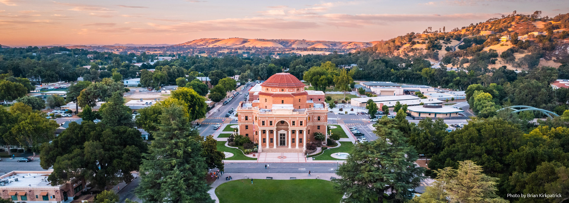 Arial shot looking down on Atascadero's Historic City Hall Building and surrounding downtown area with hills and an orange cast sunset in the background.