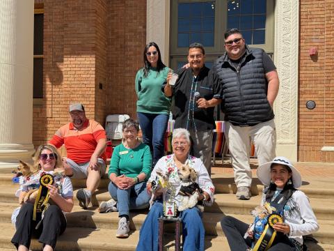 Group photo of the 2026 Pet Chihuahua Costume Contest winners seated on the steps of Atascadero City Hall with their costumed dogs, holding trophies and ribbons, with the event MC and judges sitting and standing behind them.
