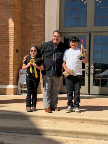 Group photo of the 2026 Tamale Festival Tamale Eating Contest winners (age 11 and under), with one person holding a trophy and another holding a ribbon, standing on either side of the event MC.