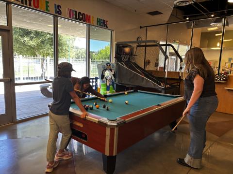 A photo of three teens gathered around a green pool table playing a game of pool.