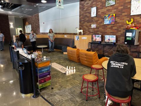 A photo of the Teen Center featuring two teens using mini bowling pins while two teens sit in large orange bean bag chairs playing on the ps5 game systems.
