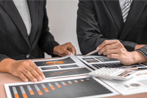 Scattered graphs on a table with one set of hands holding one of the papers and another pointing at the paper with a pen.