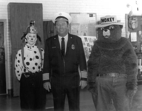 Black and white photo of Fire Chief Micheal Glenn Hicks with two people in costume on either side of him, one as a Dalmation dog and the other as Smokey the Bear.