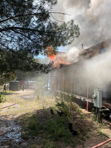 Firefighters and fire engines responding to a structure fire, with bright orange flames visible along the roofline and thick smoke rising from the building.
