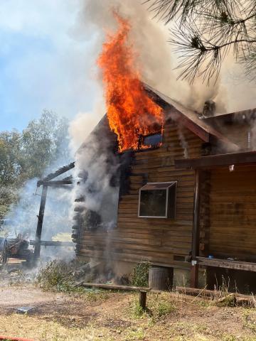 Close-up view of orange flames pouring from a second-story window during a structure fire, with large plumes of smoke rising from the building.