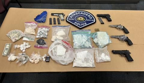 Image of items seized during the dismantling of a drug ring, neatly laid out on brown paper, under a large replica of the Atascadero Police Department badge. Items shown include a variety of bagged drugs and guns.