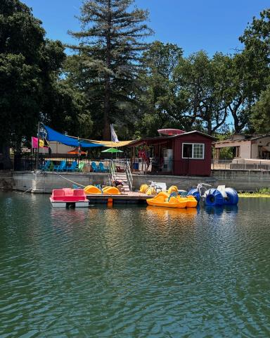 Image of paddleboats on the Atascadero Lake with small building and patio covered by colorful shade sails in the background.