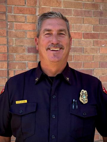 Battalion Chief David Van Son smiling in a dark blue uniform featuring a gold fire badge and nameplate.