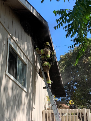 Firefighter in protective gear standing at the top of a ladder inspecting a charred roofline after a fire.