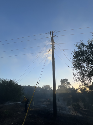 Charred ground and a damaged power pole with smoke rising as a firefighter in protective gear hoses down the area.