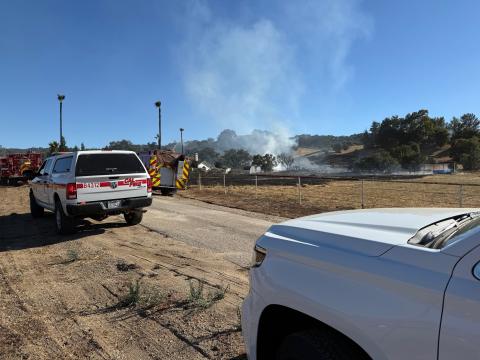 Emergency vehicles parked near residential structures, surrounded by a charred and still-smoldering field following a fire on San Carlos Rd..